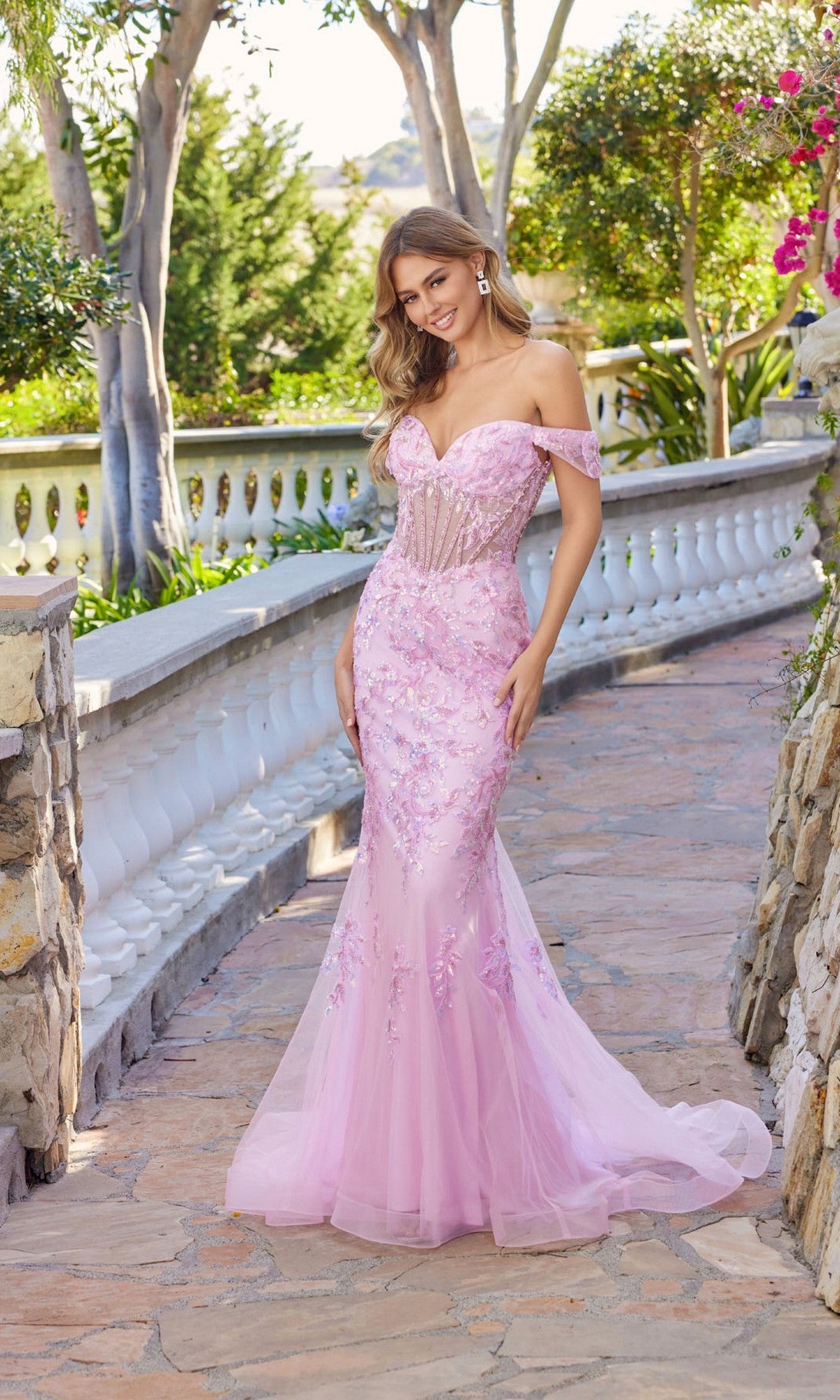 A woman in a baby pink Juliet JT2612N long prom dress with off-the-shoulder mermaid silhouette, sheer corset bodice, and floral appliqué stands on a stone walkway in a garden.